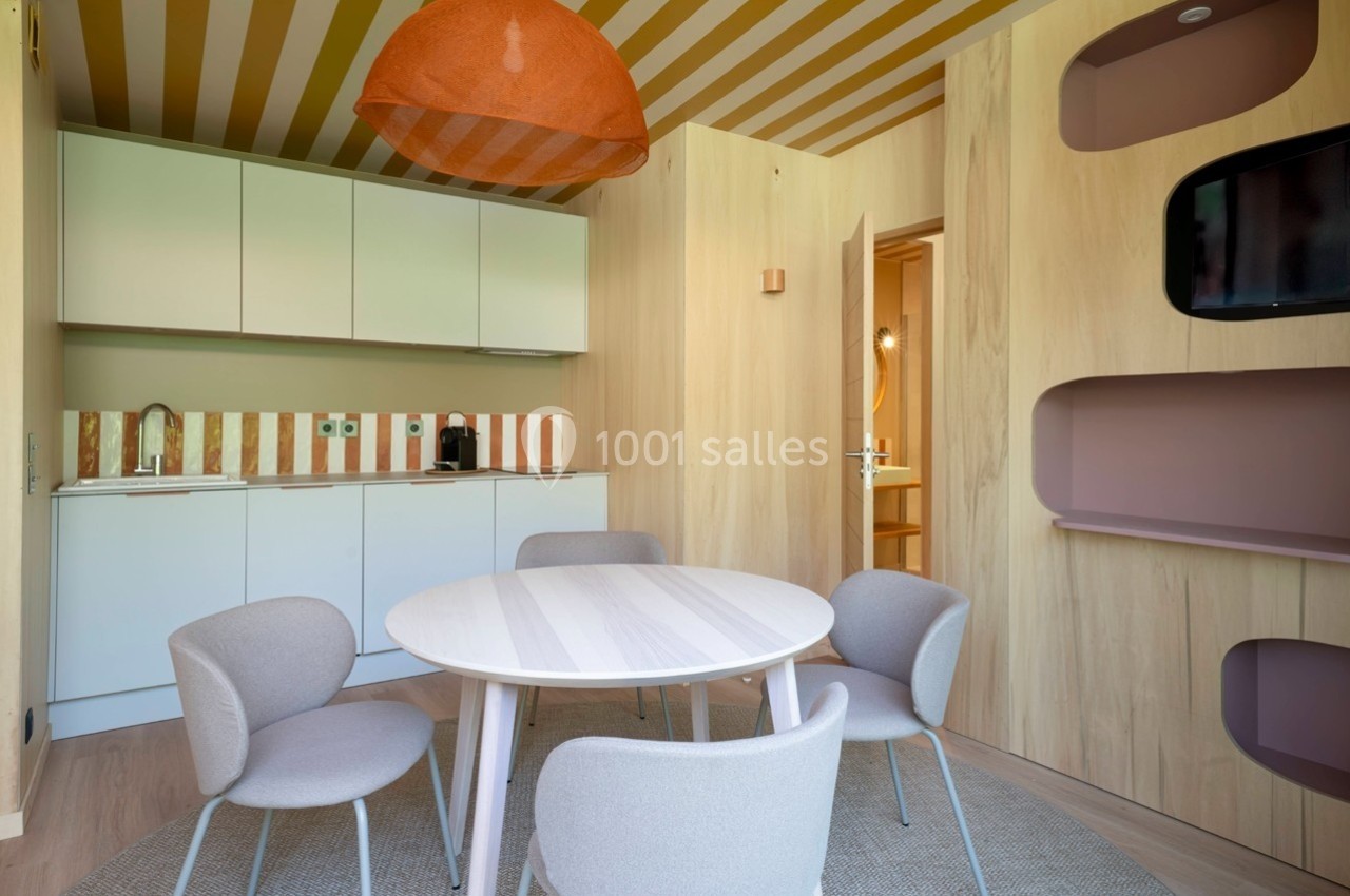 Salle à manger moderne avec table ronde, chaises grises, cuisine équipée en arrière-plan et éclairage suspendu orange.