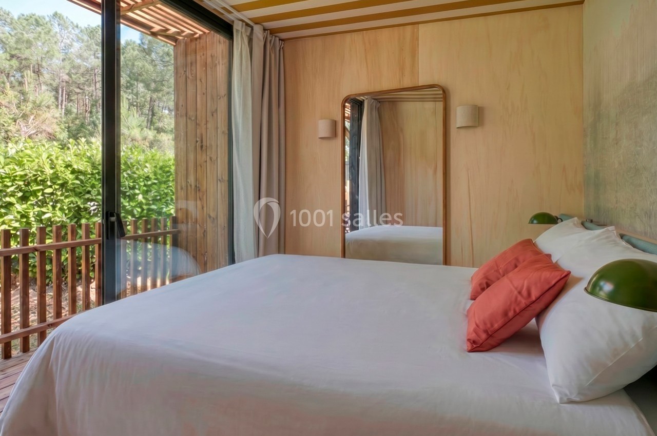 Chambre lumineuse avec lit double, miroir mural, grandes baies vitrées et vue sur une terrasse en bois et des arbres.