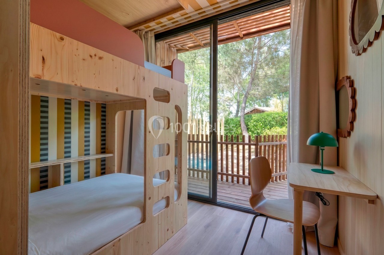 Chambre en bois avec lits superposés, bureau, et vue sur une terrasse donnant sur un jardin arboré.