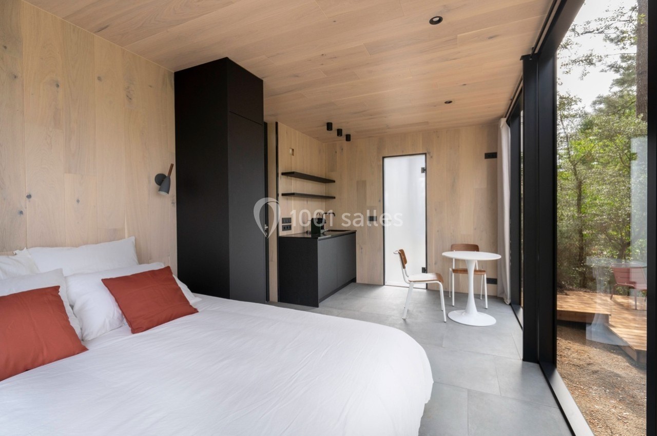 Intérieur d'une petite maison moderne avec lit, cuisine minimaliste, table ronde et grande baie vitrée donnant sur la nature.