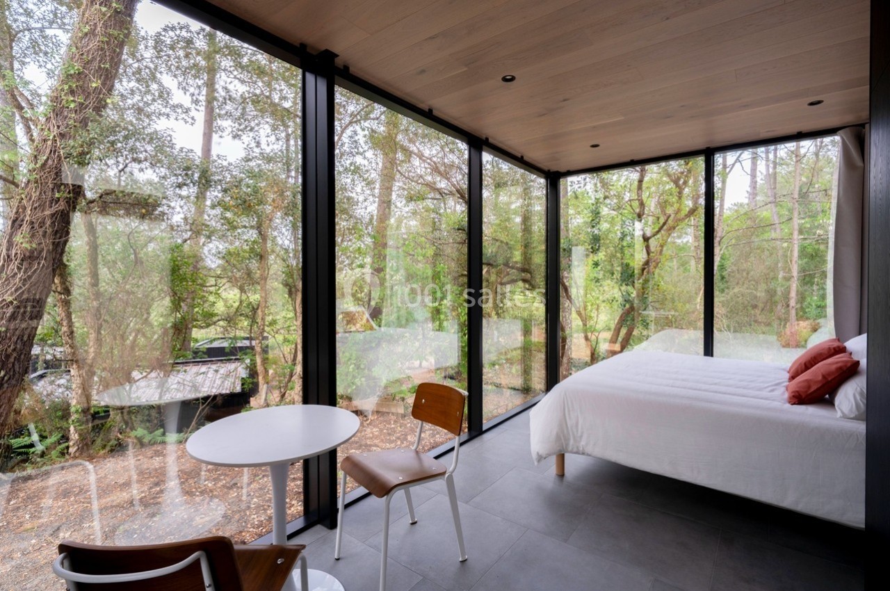 Chambre moderne avec grandes baies vitrées offrant une vue sur une forêt, table et chaises en bois clair.