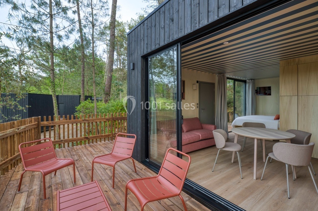 Terrasse en bois avec chaises rouges, donnant sur un intérieur moderne avec salon, table et lit, entourée de verdure.
