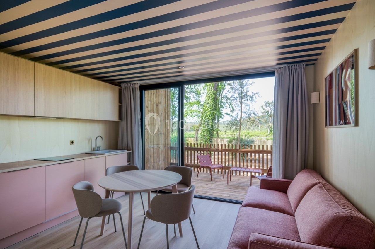 Intérieur d'une pièce lumineuse avec cuisine, table, canapé et accès à une terrasse en bois donnant sur un espace verdoyant.