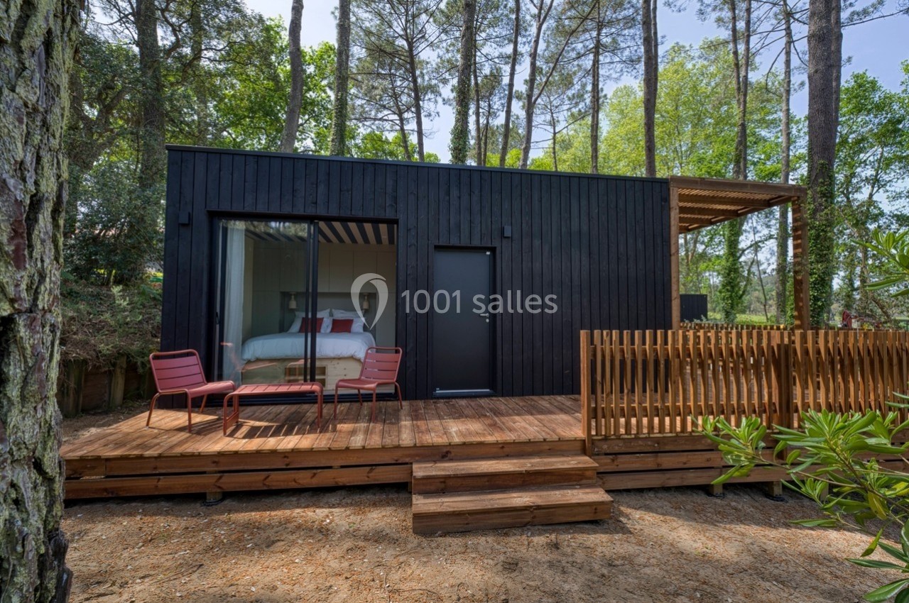 Cabane moderne en bois noir avec terrasse, entourée d'arbres, équipée de chaises rouges et d'une table basse.