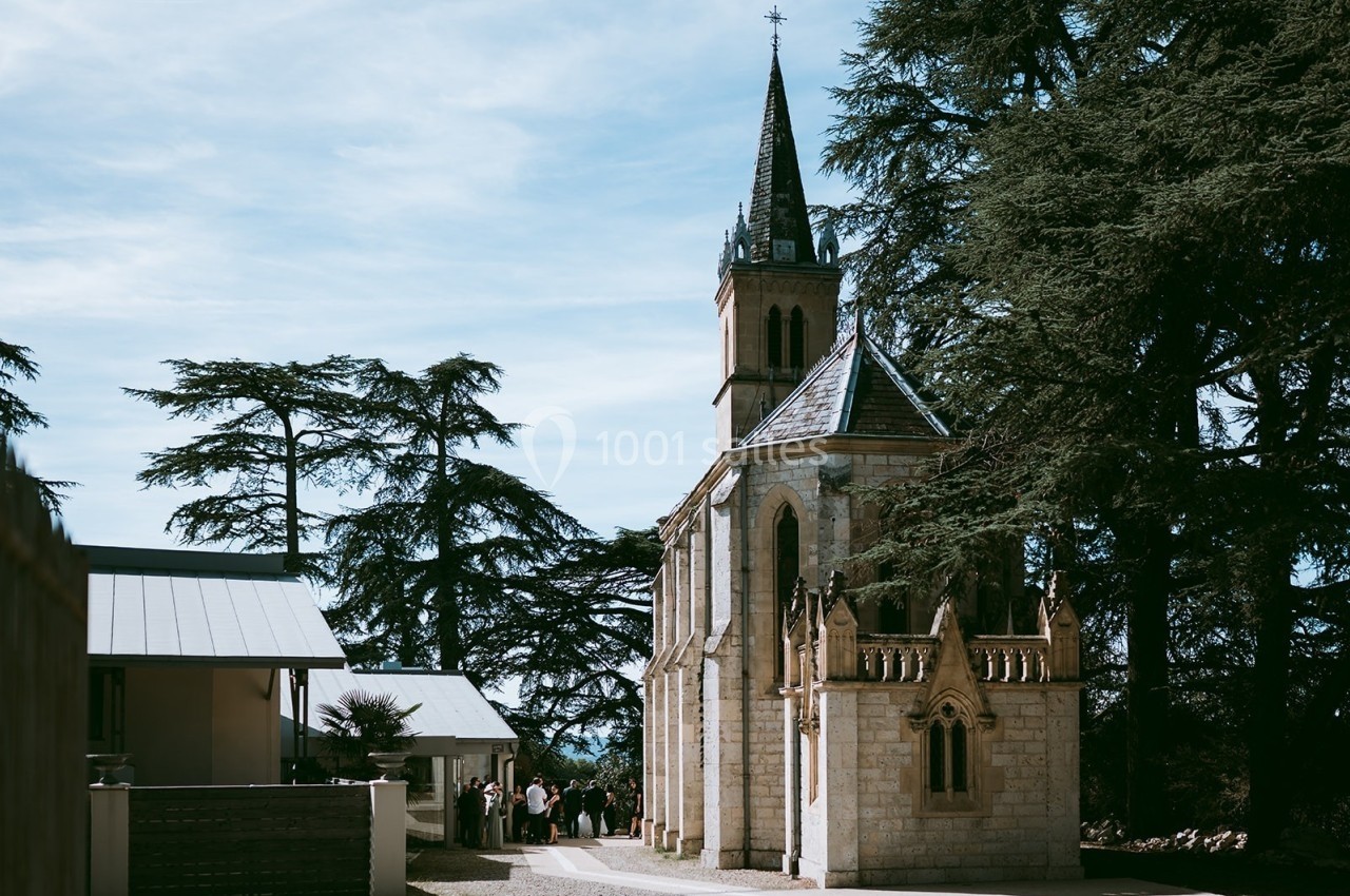 Église en pierre avec clocher entourée d'arbres, des personnes rassemblées à proximité sous un ciel dégagé.