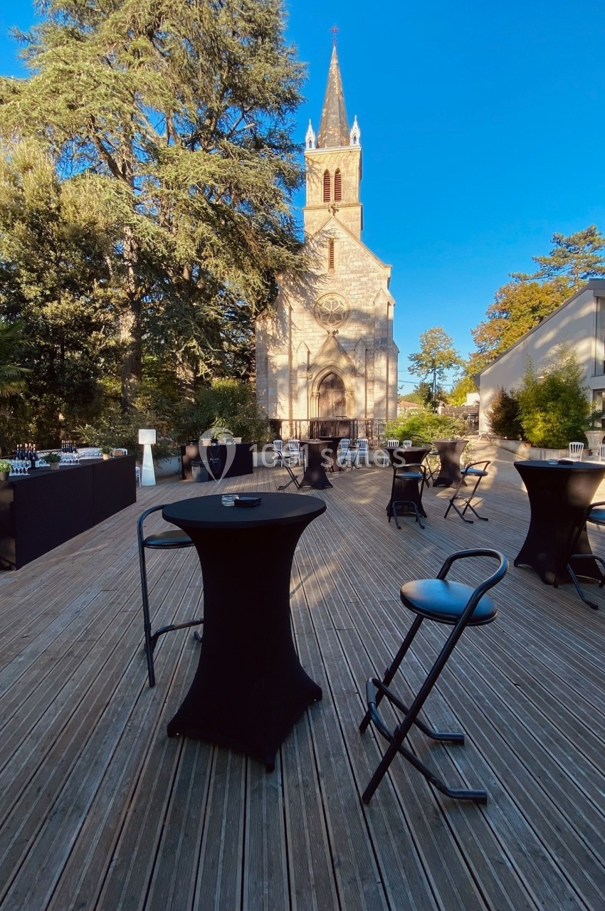 Terrasse en bois aménagée avec tables hautes et chaises, vue sur une église entourée de végétation sous un ciel bleu.