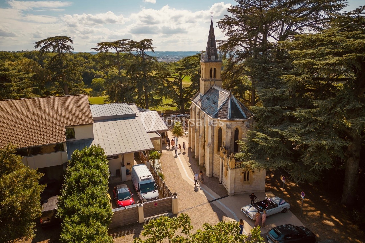 Vue aérienne d'une petite chapelle entourée d'arbres, avec des bâtiments modernes et des voitures à proximité.