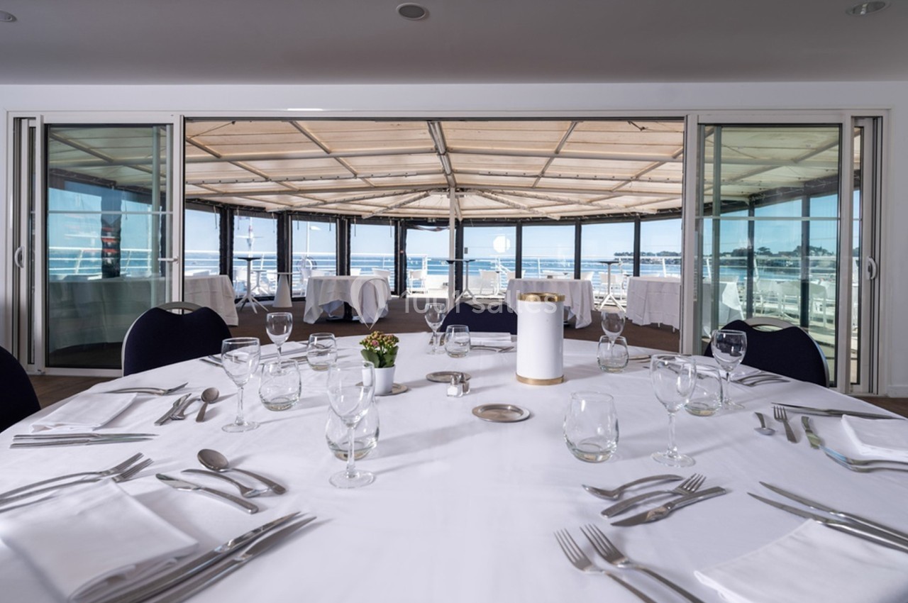 Table dressée avec vaisselle et verres, située dans une salle lumineuse avec vue sur une terrasse et la mer.