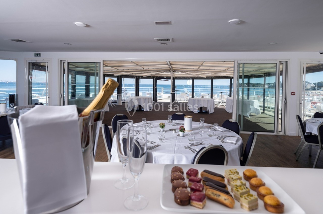 Salle de réception lumineuse avec vue sur la mer, tables dressées et plateau de desserts au premier plan.