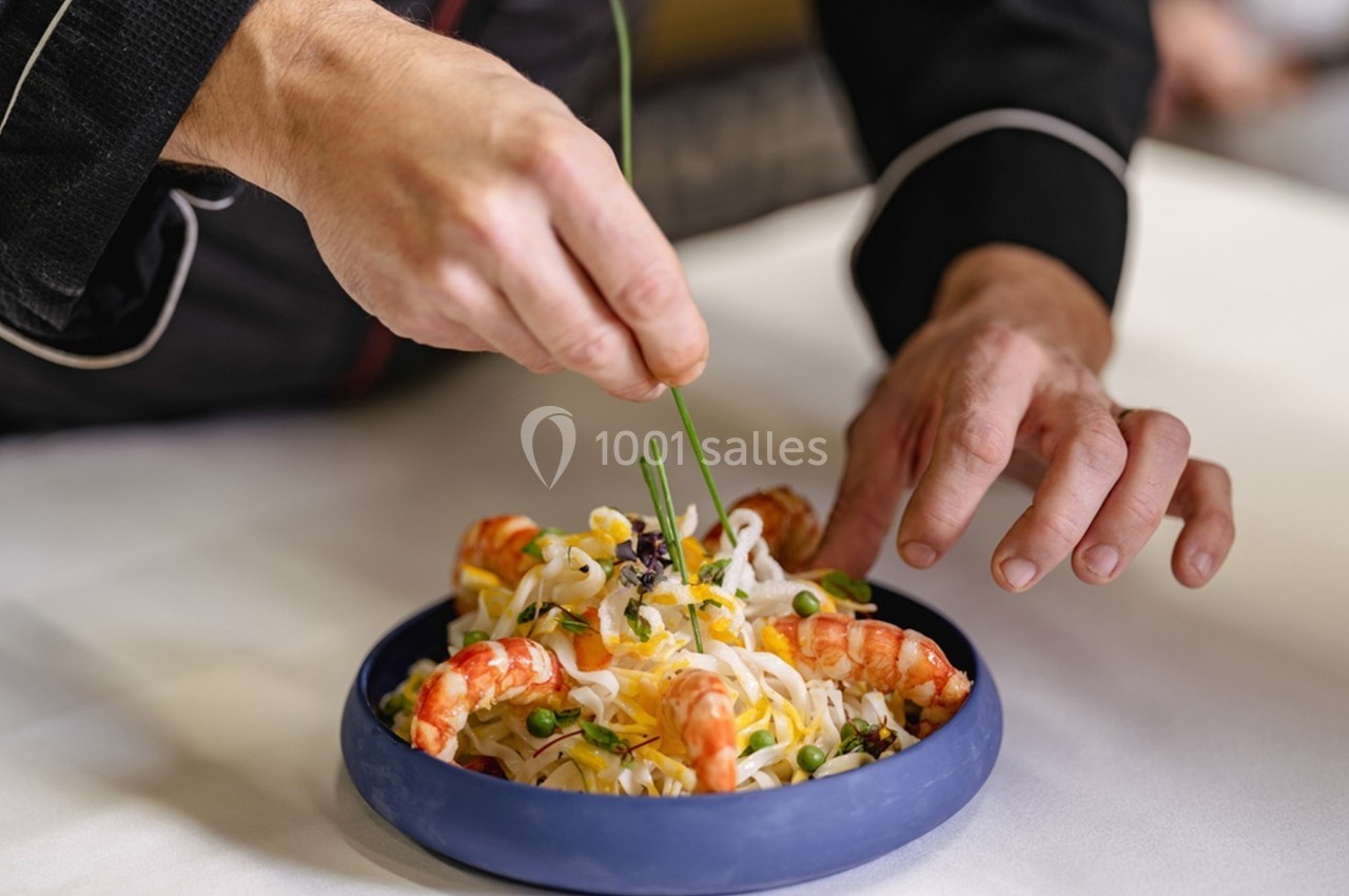 Un chef décore un plat de nouilles aux crevettes et légumes avec des brins de ciboulette.