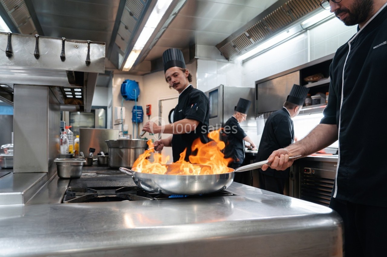 Cuisiniers travaillant dans une cuisine professionnelle, avec une poêle enflammée au premier plan.