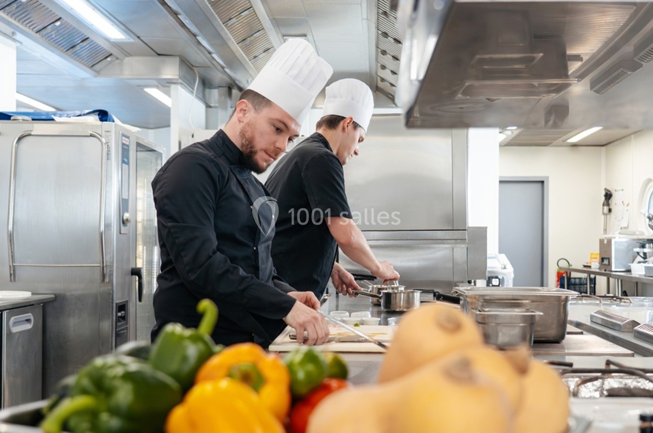 Deux chefs cuisiniers en tenue travaillent dans une cuisine professionnelle avec des légumes au premier plan.