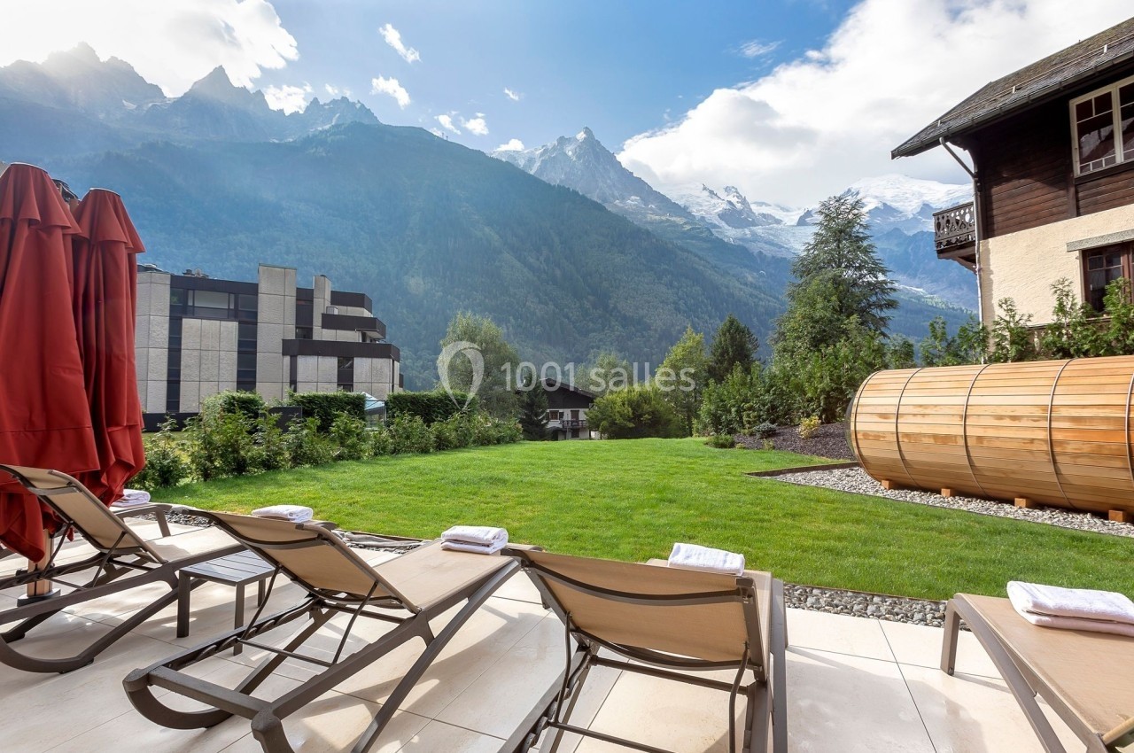 Chaises longues alignées sur une terrasse avec vue sur un jardin, des montagnes enneigées et un sauna en bois.
