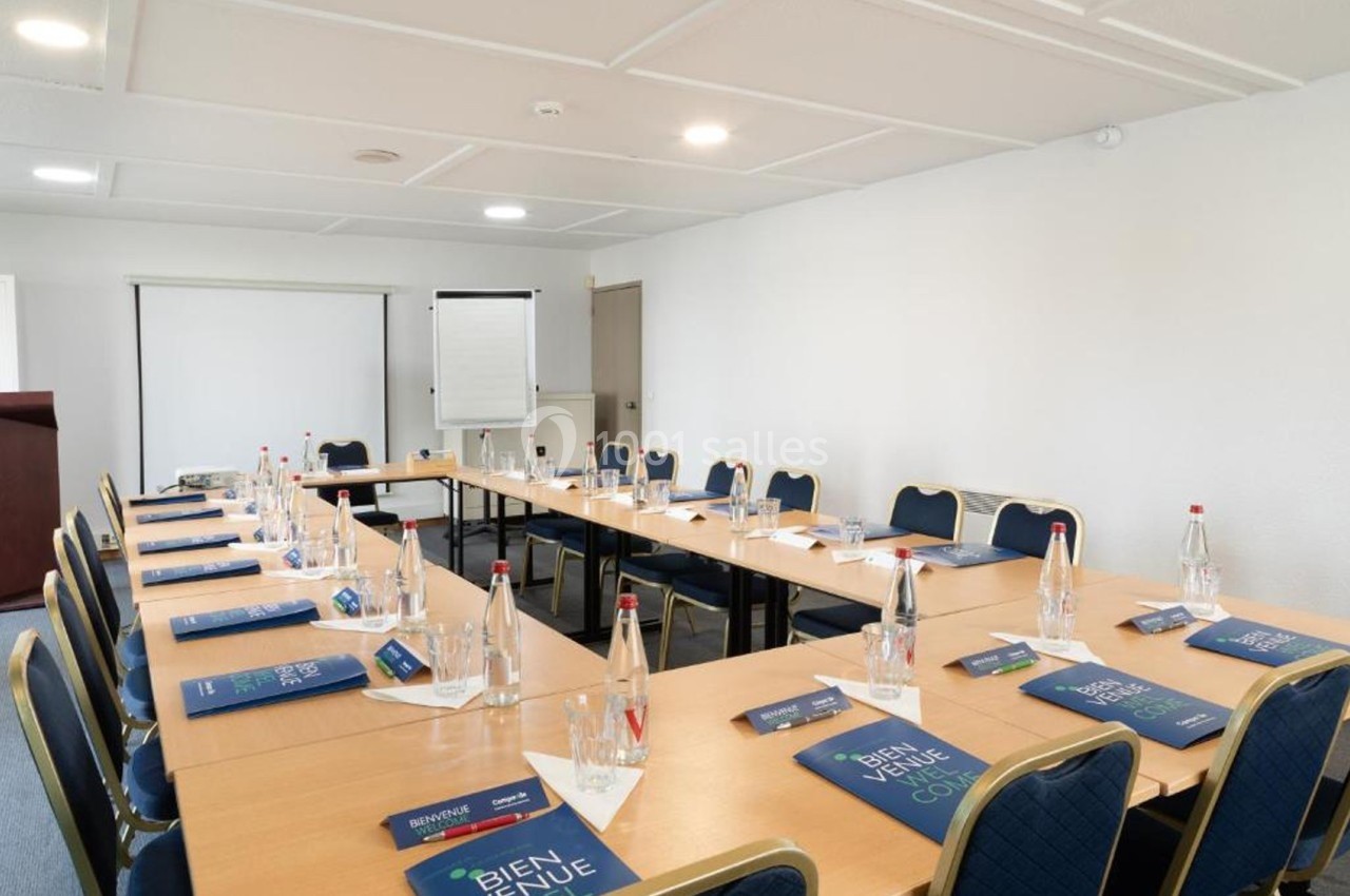 Salle de réunion lumineuse avec tables disposées en U, chaises, bouteilles d'eau, bloc-notes et paperboard.