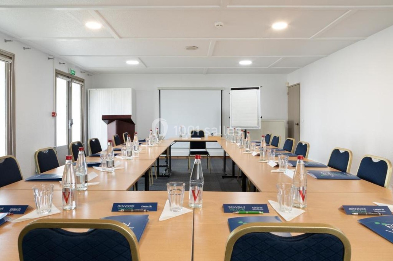 Salle de réunion lumineuse avec tables disposées en U, chaises, bouteilles d'eau, paperboard et pupitre en arrière-plan.