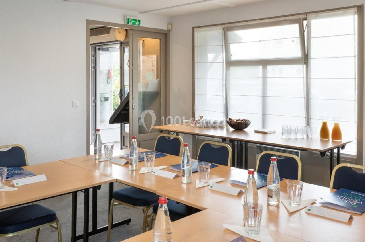 Salle de réunion lumineuse avec tables en U, chaises, bouteilles d'eau, verres, blocs-notes et carafes de jus.