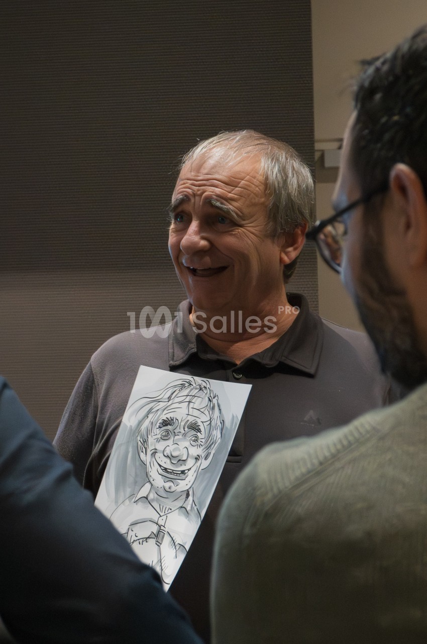 Un homme souriant tient un dessin humoristique de lui-même, entouré de personnes dans une pièce.