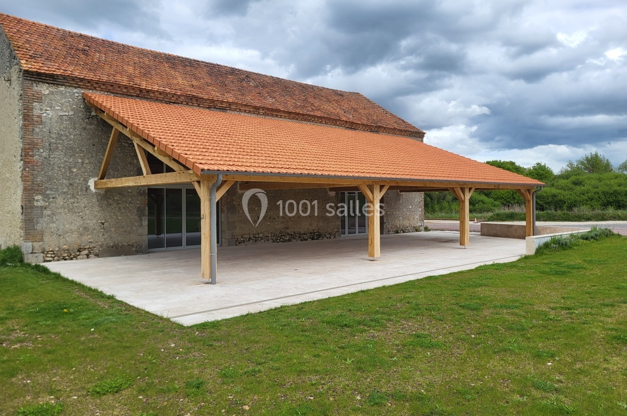 Abri en bois avec toit en tuiles rouges adossé à un bâtiment en pierre, sur une dalle en béton, entouré de verdure.