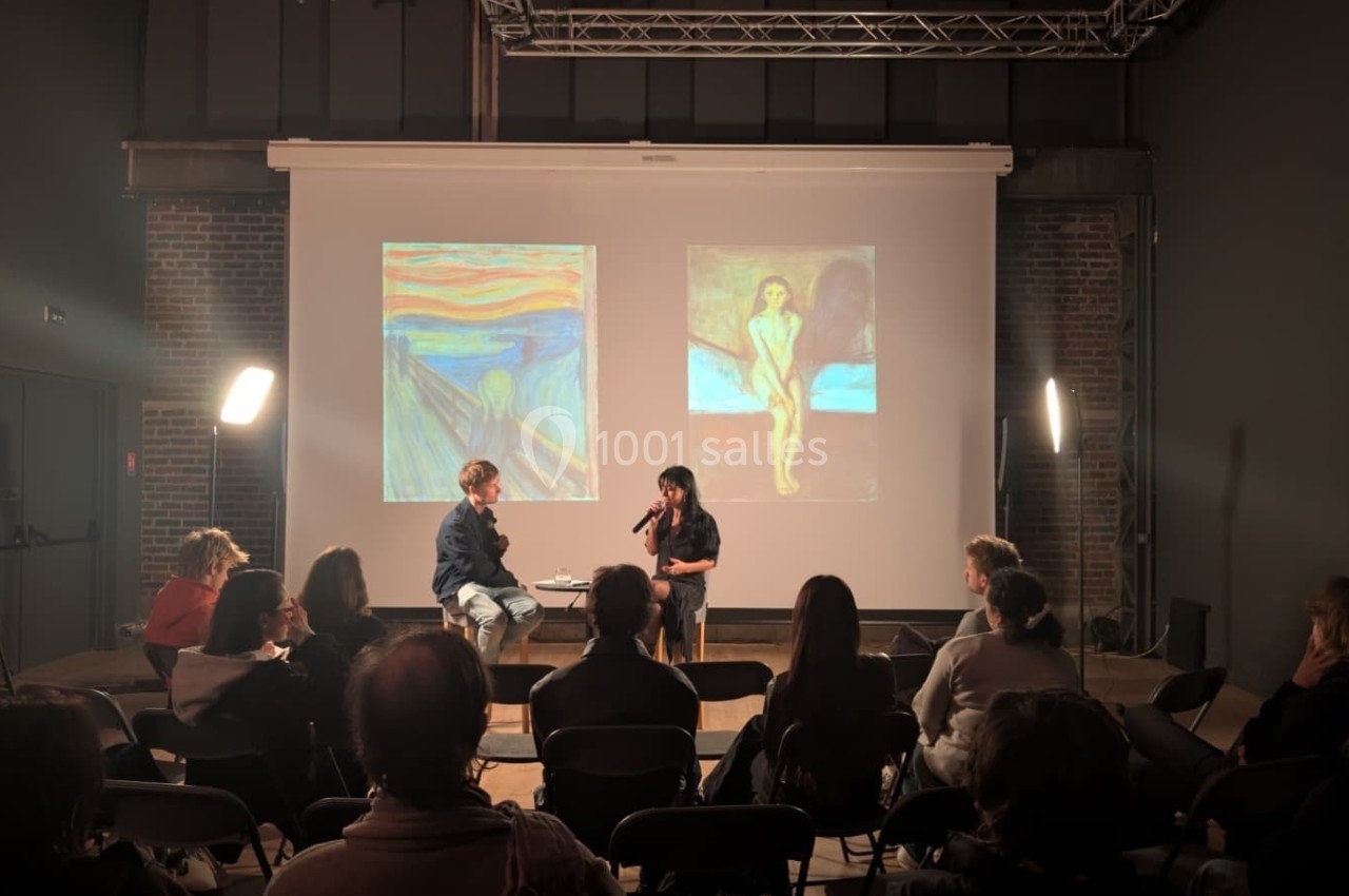 Deux personnes discutent sur une scène devant un public, avec des œuvres d'art projetées en arrière-plan.