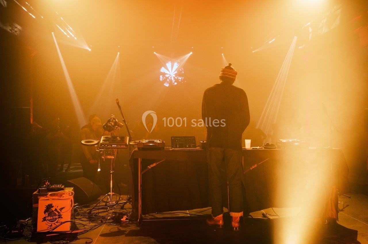 Un DJ de dos mixe sur une scène éclairée par des lumières chaudes, face à un public dans une ambiance tamisée.