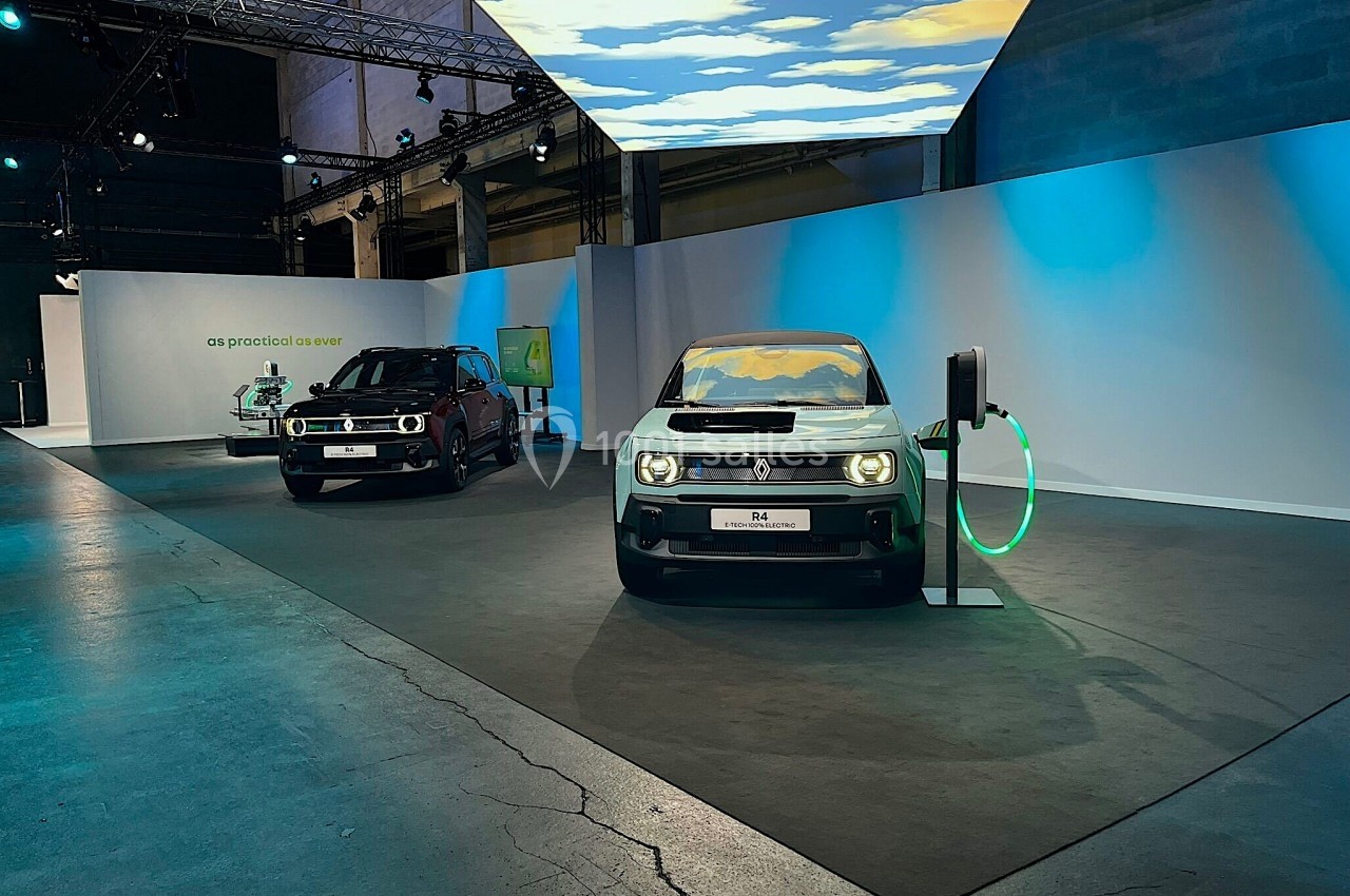 Deux voitures électriques exposées dans un espace intérieur éclairé, l'une connectée à une borne de recharge.