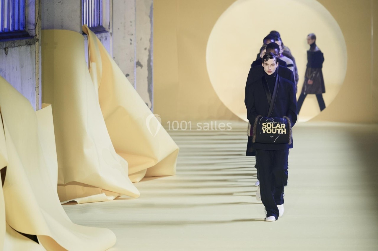 Un défilé de mode avec des mannequins en tenues sombres marchant sur un podium jaune, décor minimaliste.