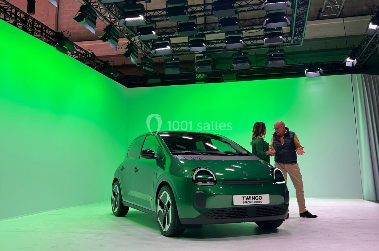 Une voiture citadine verte exposée dans un studio éclairé, avec deux personnes discutant à côté.