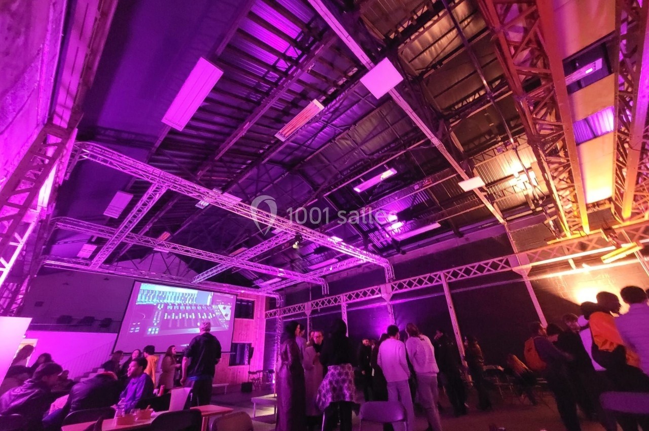 Salle industrielle avec éclairage violet, des personnes discutent et un écran projette des informations.