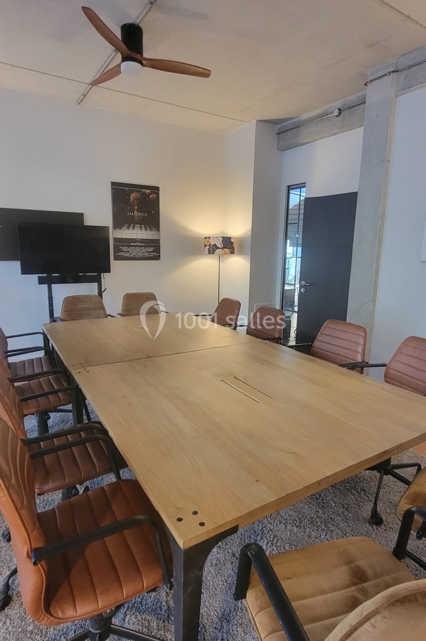 Salle de réunion avec une grande table en bois, huit chaises en cuir marron, écran mural et éclairage moderne.