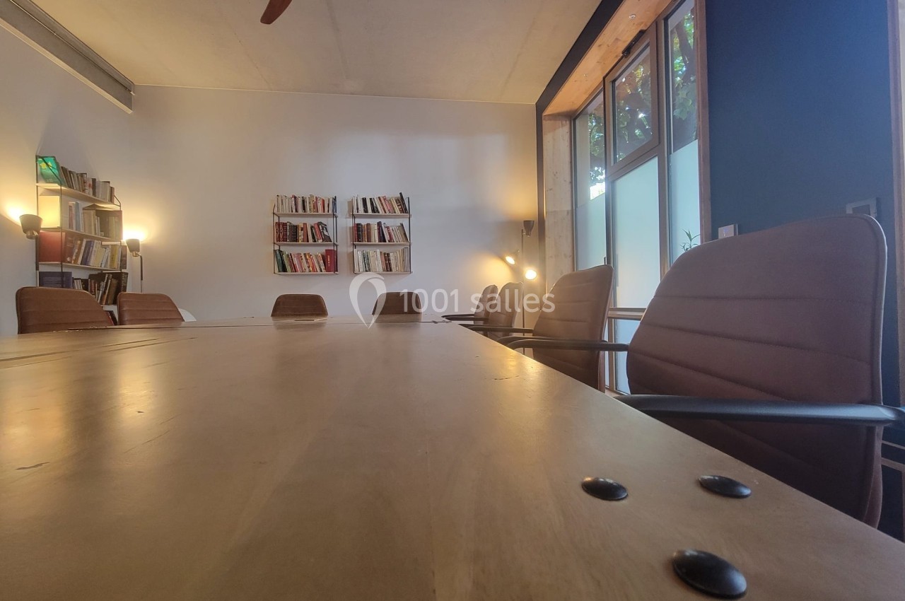 Salle de réunion avec une grande table en bois, chaises en cuir, étagères de livres et lumière naturelle.