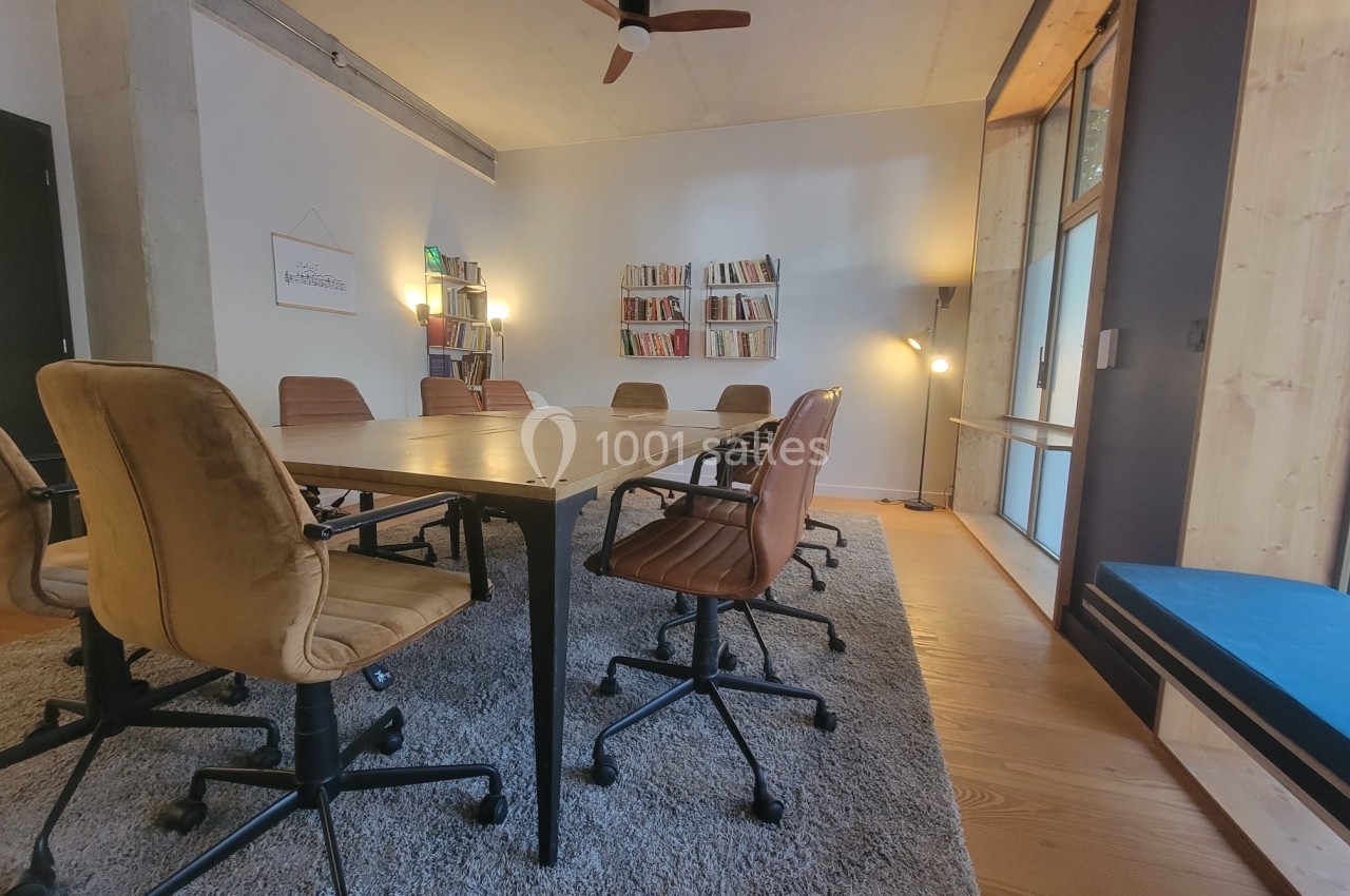 Salle de réunion lumineuse avec table en bois, chaises rembourrées, étagères de livres et grandes fenêtres.