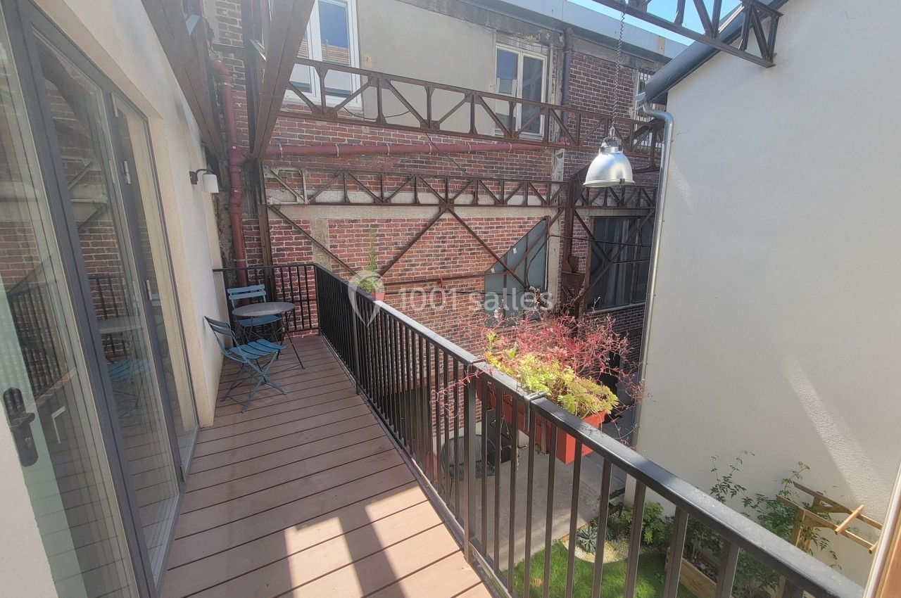 Balcon en bois avec table et chaises, surplombant une petite cour intérieure avec des murs en briques et des plantes.