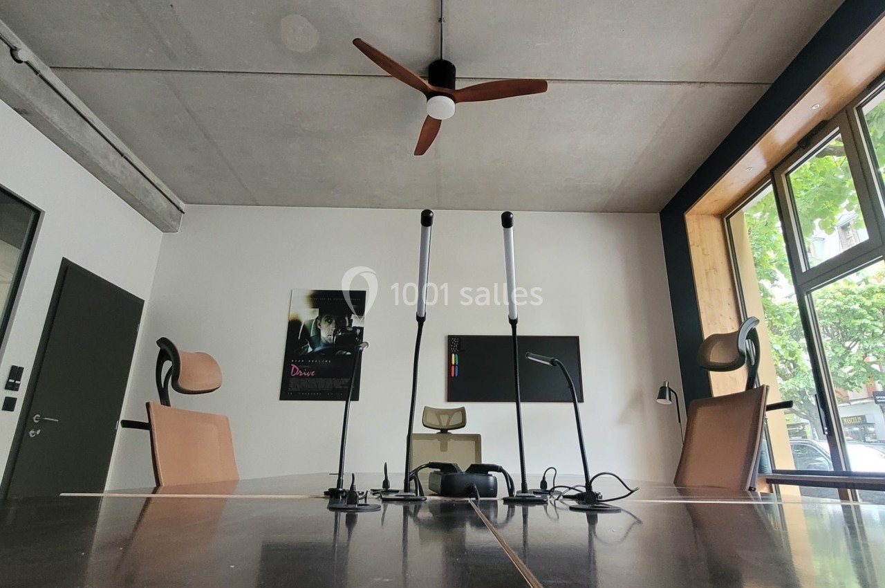 Salle de réunion moderne avec table en bois, chaises design, ventilateur au plafond et grande fenêtre donnant sur des arbres.