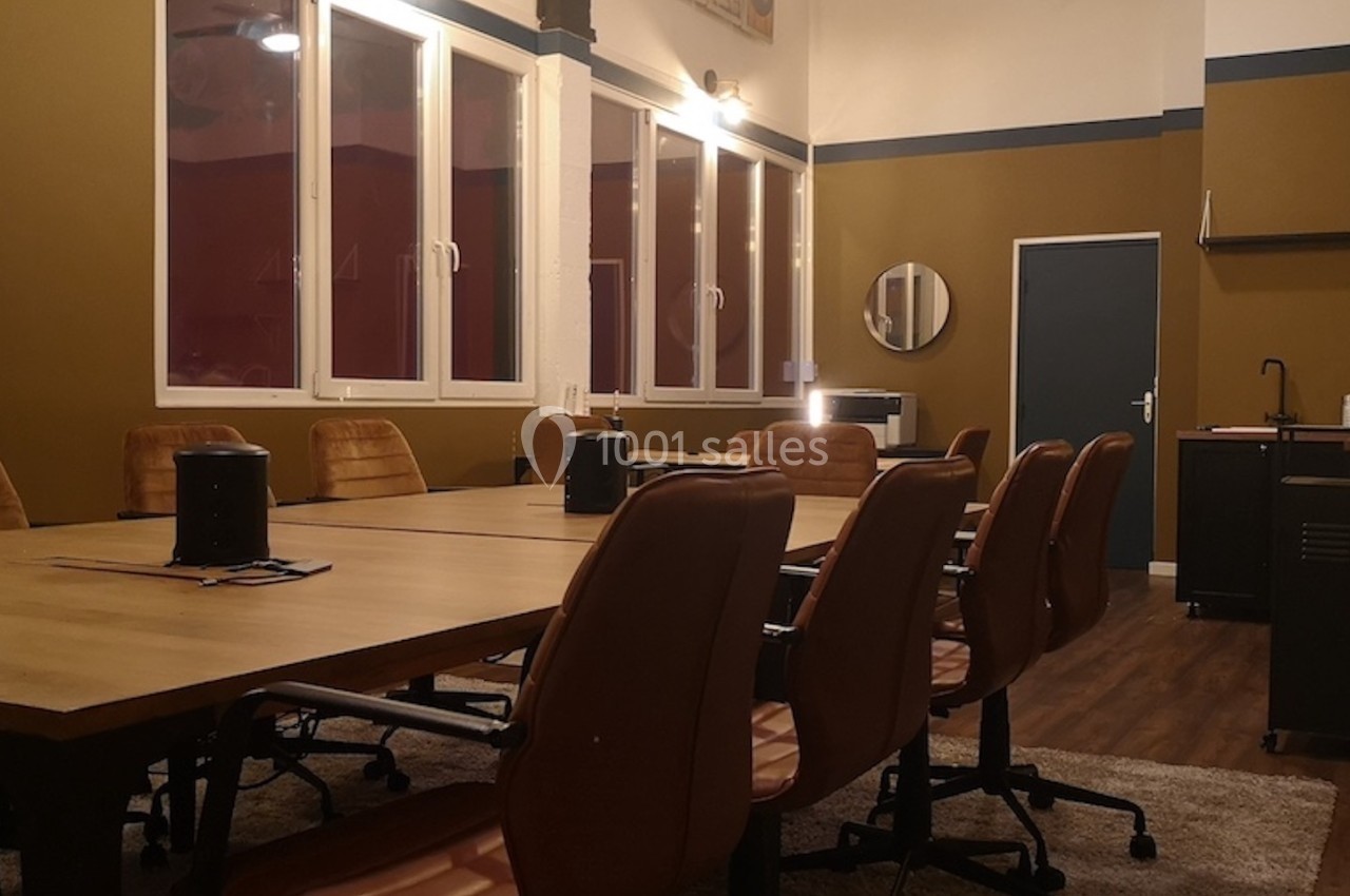 Salle de réunion avec une grande table en bois, chaises en cuir marron, fenêtres et kitchenette en arrière-plan.