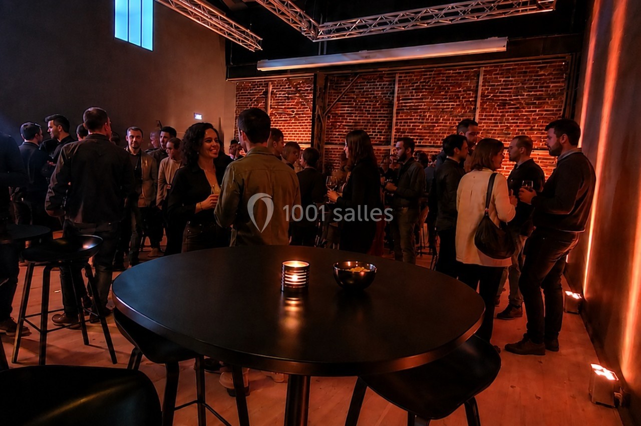 Groupe de personnes discutant dans une salle avec éclairage tamisé, murs en briques et tables hautes.