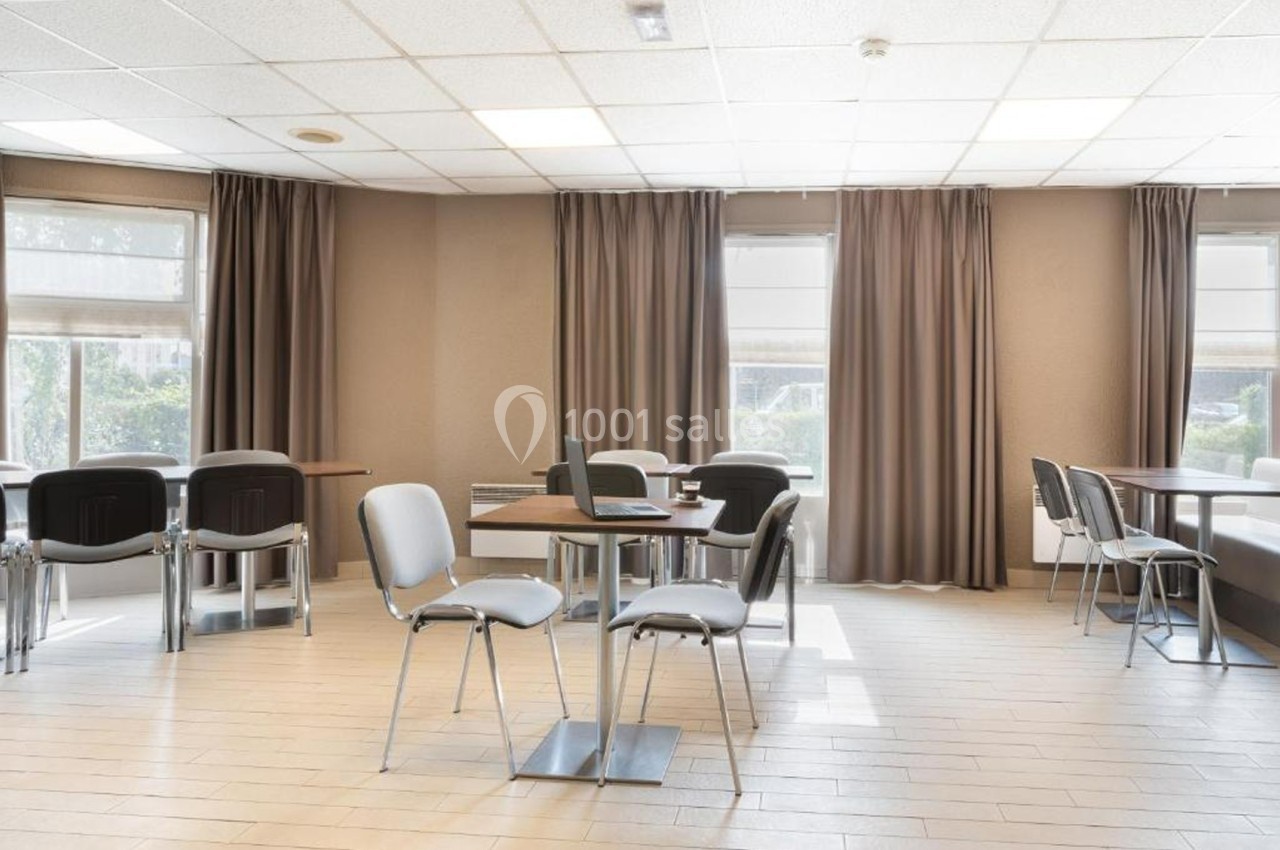 Salle lumineuse avec tables, chaises et rideaux, aménagée pour des réunions ou des espaces de travail.
