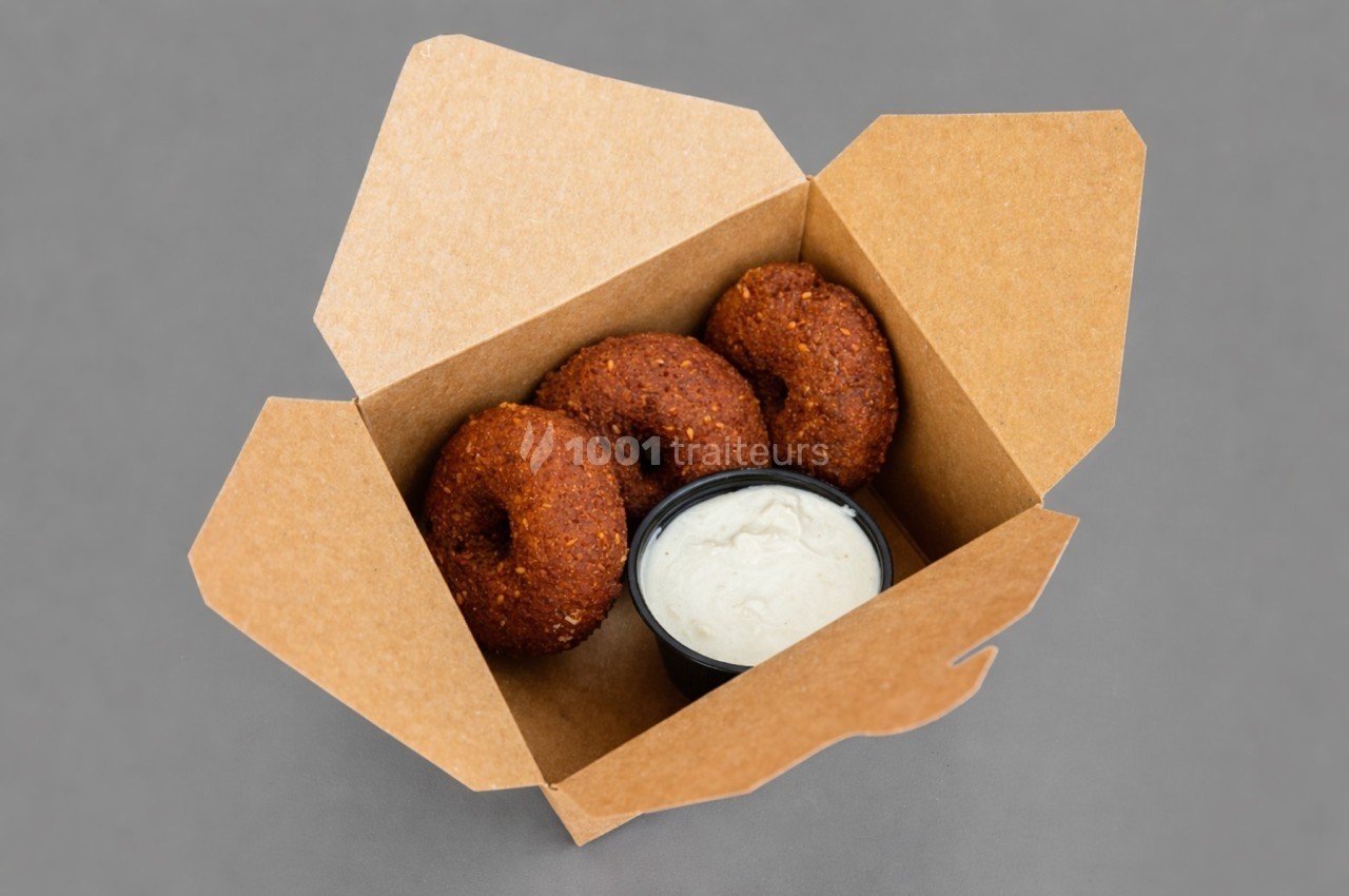 Trois falafels ronds dans une boîte en carton avec une sauce blanche dans un petit récipient noir.