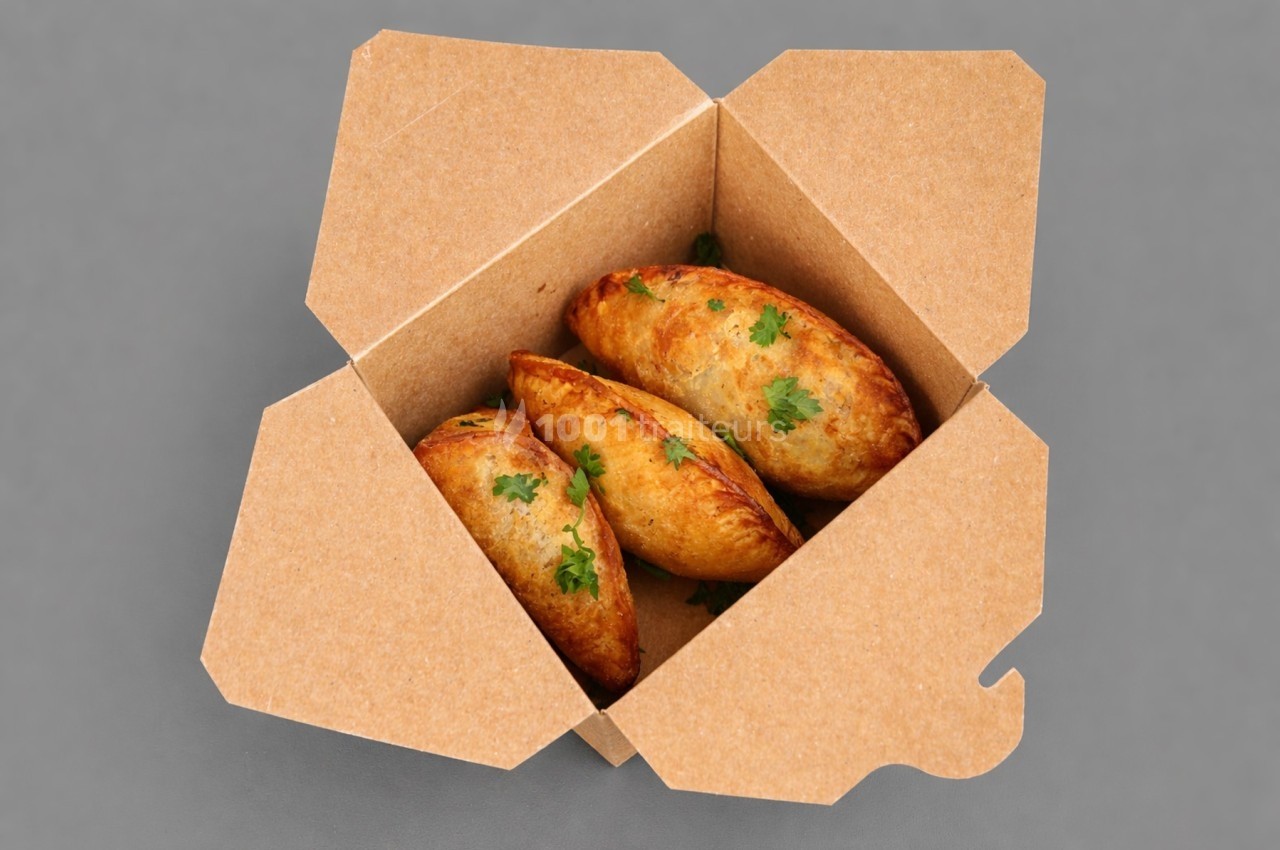 Trois empanadas dorées garnies de persil frais dans une boîte en carton ouverte sur fond gris.