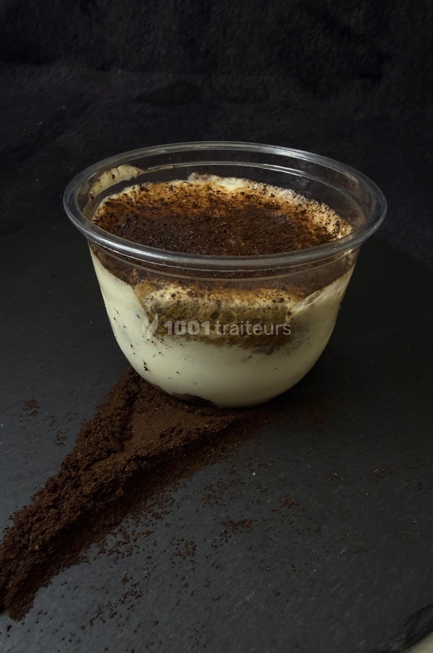 Dessert tiramisu dans un pot transparent, posé sur une surface sombre avec de la poudre de cacao répandue.