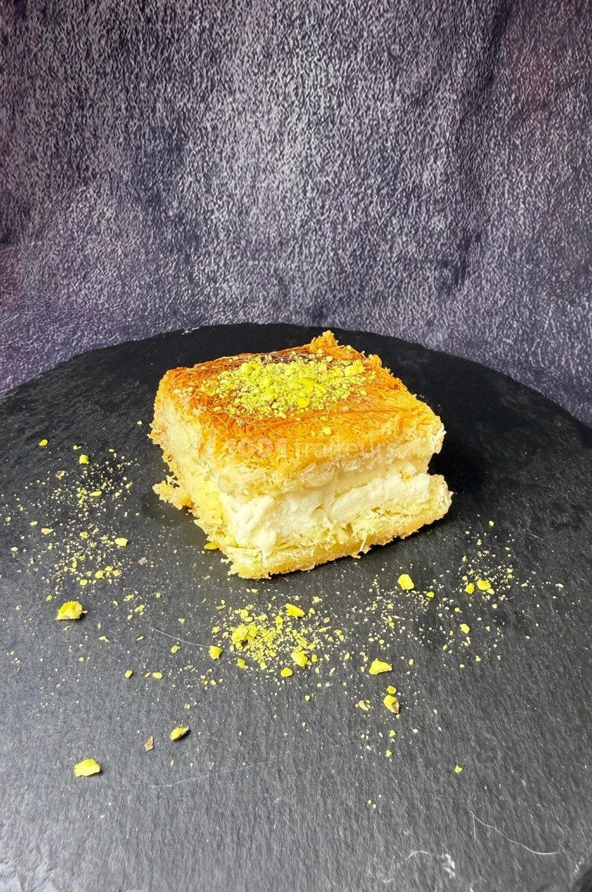 Part de gâteau à base de pâte croustillante, garnie de crème et parsemée de pistaches sur une assiette noire.