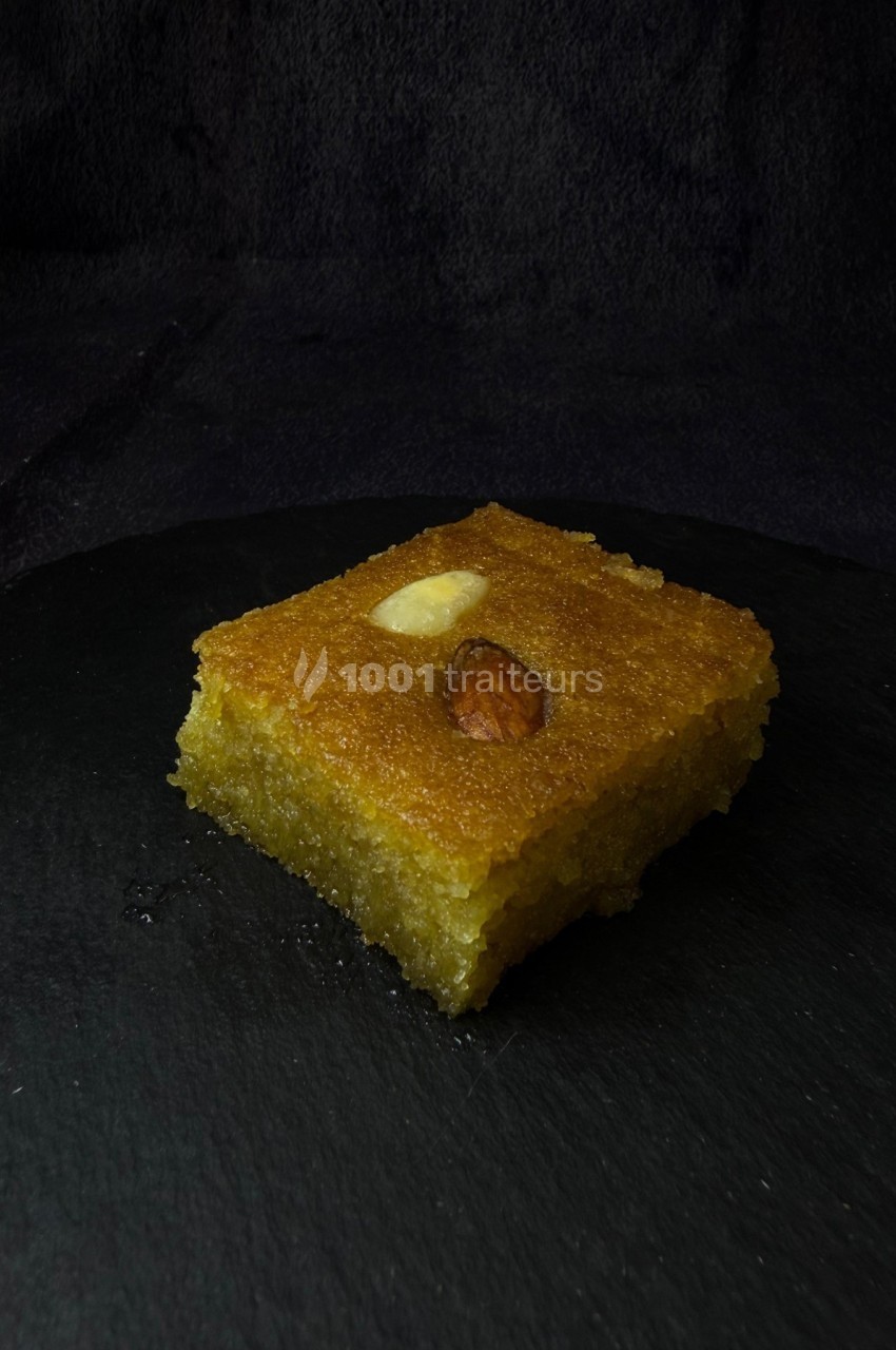 Part de gâteau oriental doré, imbibé de sirop, garnie d'une amande et posée sur une surface noire.