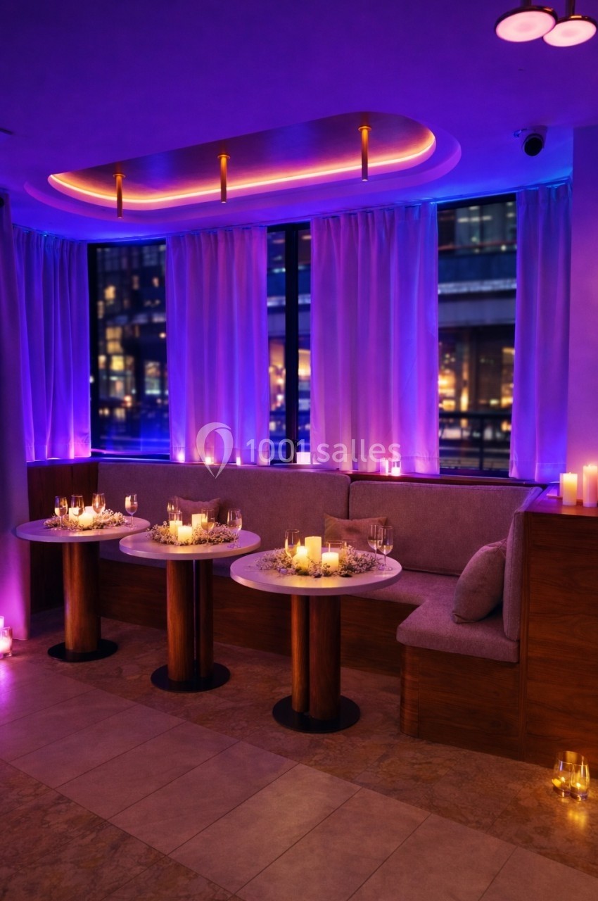 Salle élégante avec tables décorées de bougies et fleurs, éclairage tamisé violet et grandes fenêtres donnant sur la ville.