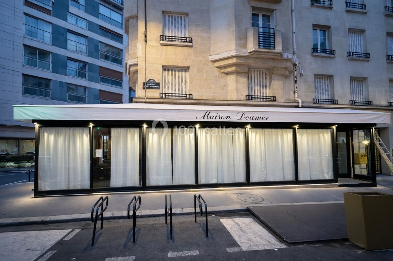 Façade d'un restaurant avec de grandes baies vitrées et rideaux blancs, situé au rez-de-chaussée d'un immeuble urbain.