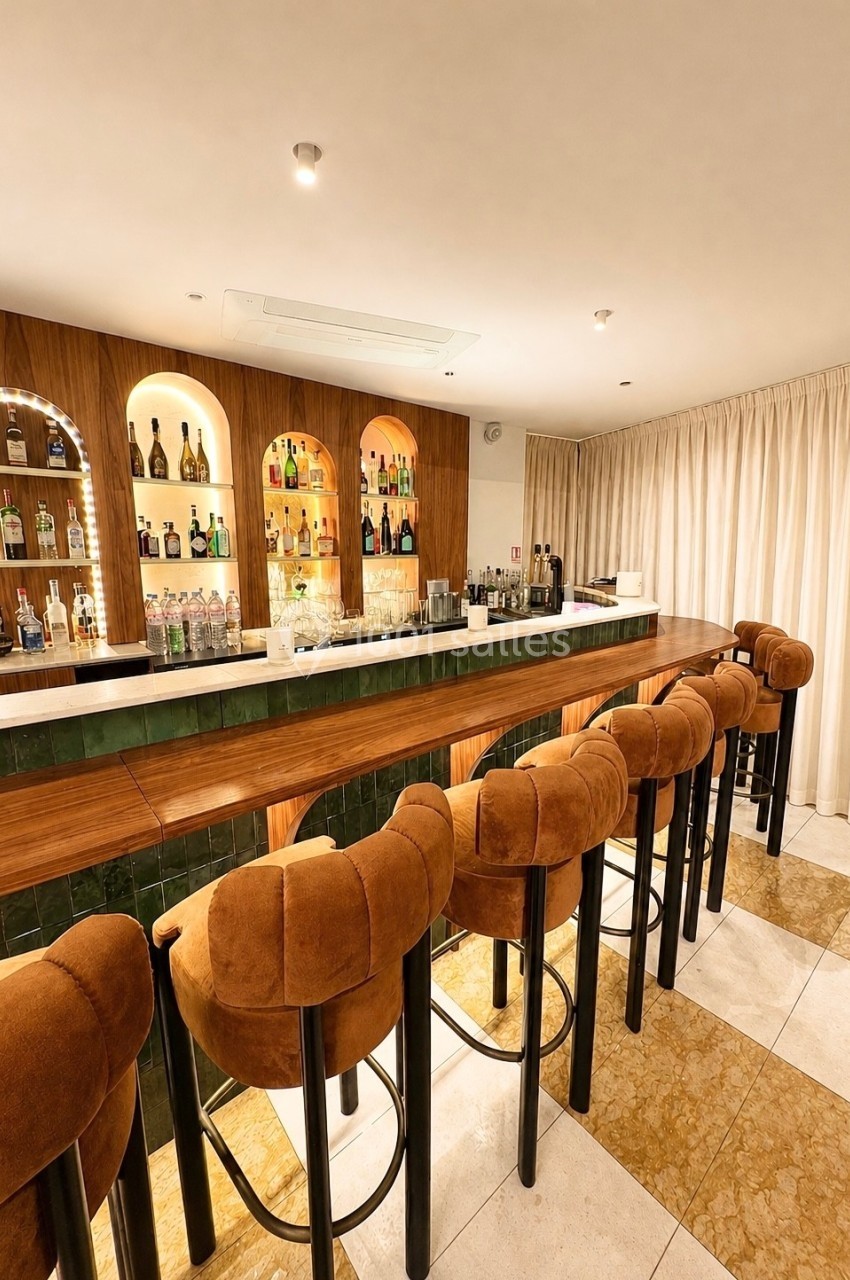 Bar élégant avec comptoir en bois, tabourets rembourrés et étagères illuminées garnies de bouteilles.
