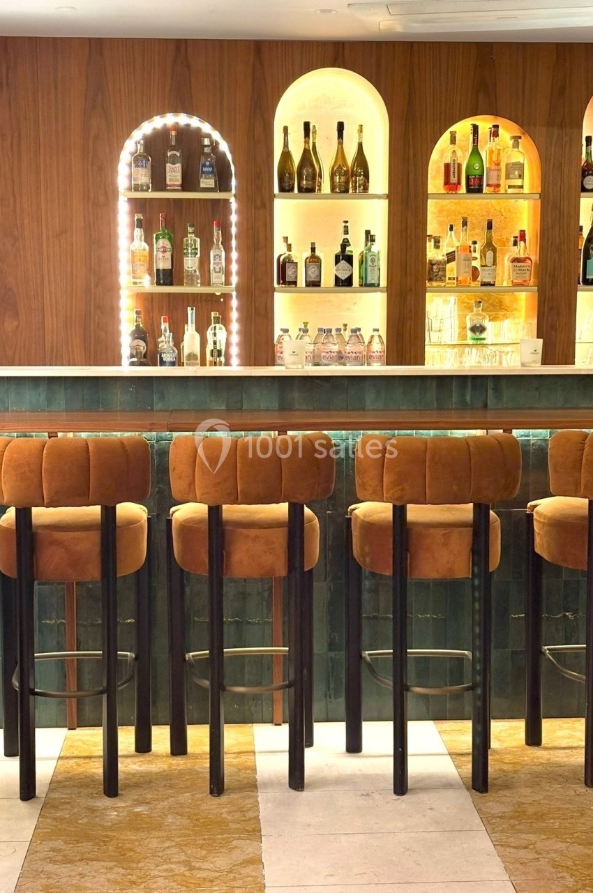 Bar avec comptoir en marbre, tabourets en velours orange et étagères éclairées exposant des bouteilles d'alcool.