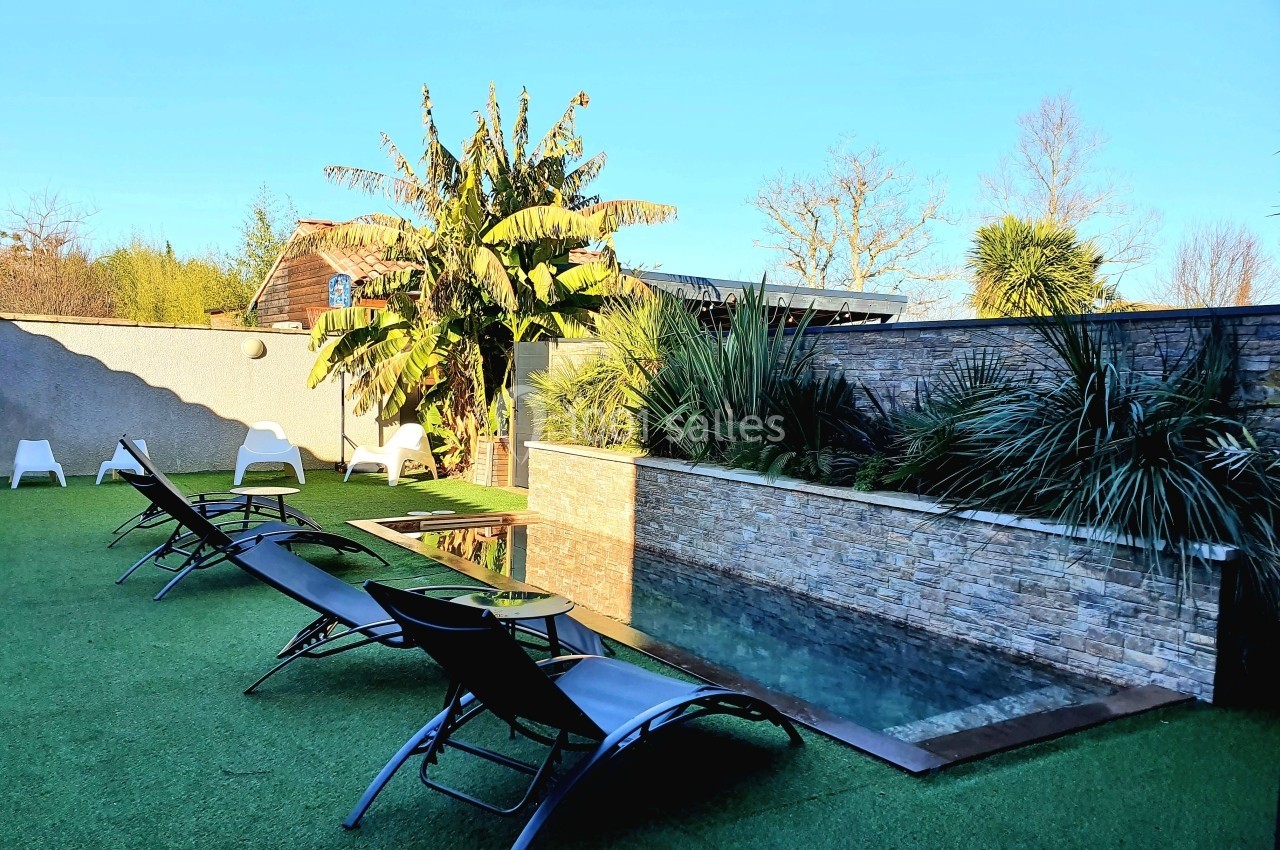 Piscine extérieure entourée de transats noirs, végétation tropicale et mur en pierre dans un jardin ensoleillé.