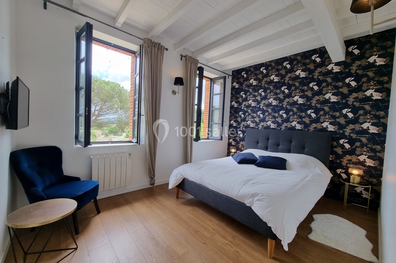Chambre lumineuse avec lit double, fauteuil bleu, murs blancs et papier peint à motifs sombres, vue sur l'extérieur.