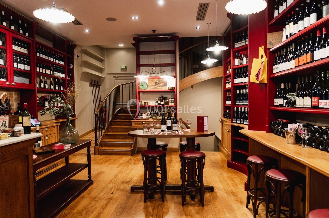 Intérieur d'une cave à vin avec étagères remplies de bouteilles, table centrale et escalier en bois au fond.