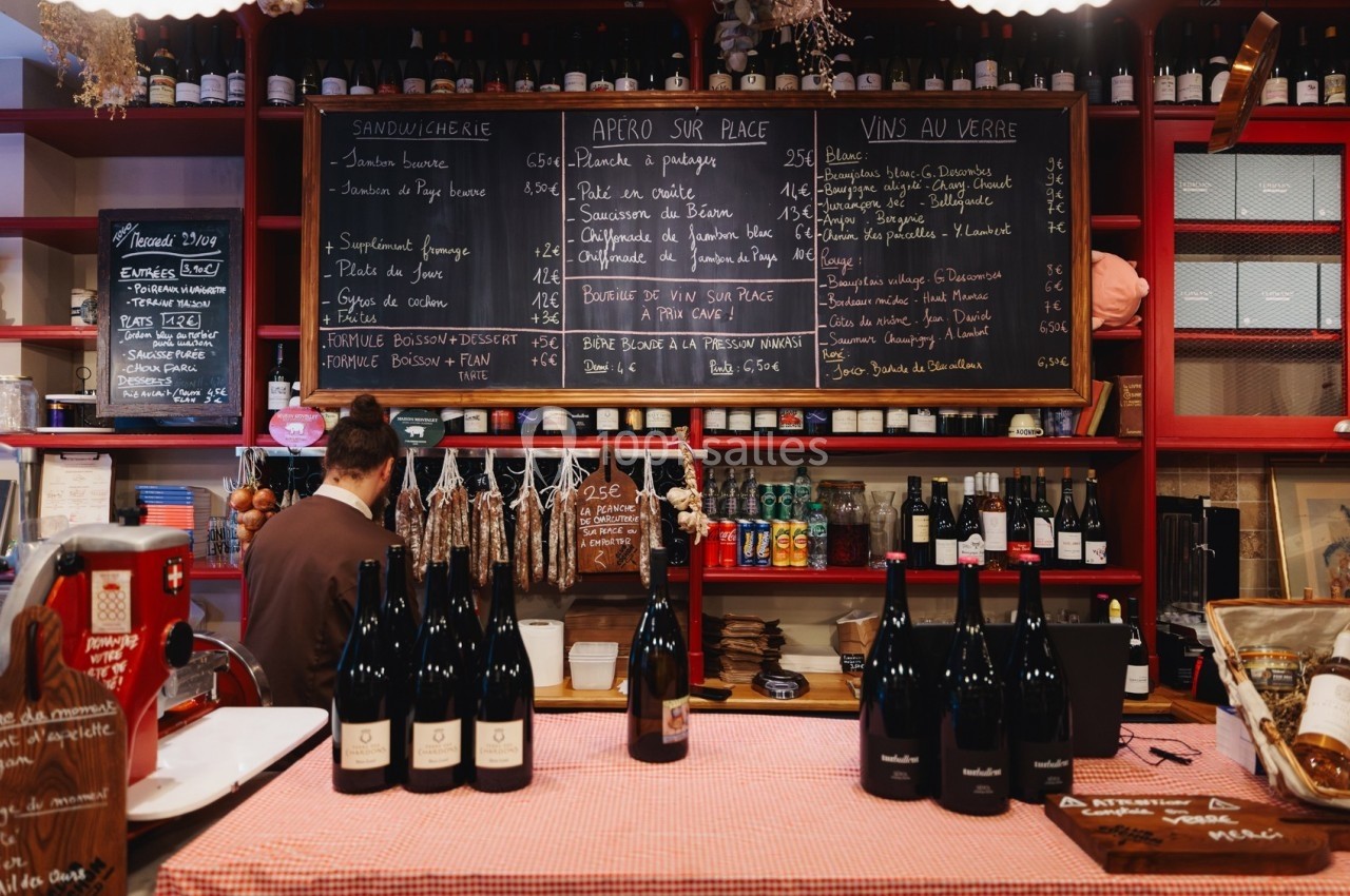 Comptoir d'une épicerie avec bouteilles de vin, saucissons suspendus et ardoises affichant menus et prix.
