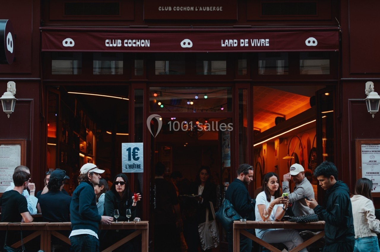 Façade d'un bar animé avec des clients attablés en terrasse, éclairage chaleureux à l'intérieur.